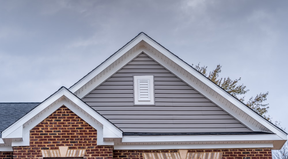 White attic gable vent louver, white square window, triangle with red brick or gray beige horizontal lap vinyl siding on a new construction luxury american single family home in the East Coast USA.
