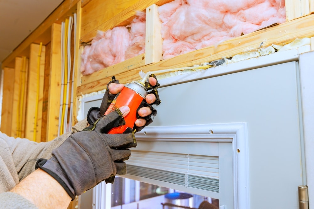 Person applies weatherproofing foam spray around door frame during home renovation residential setting.