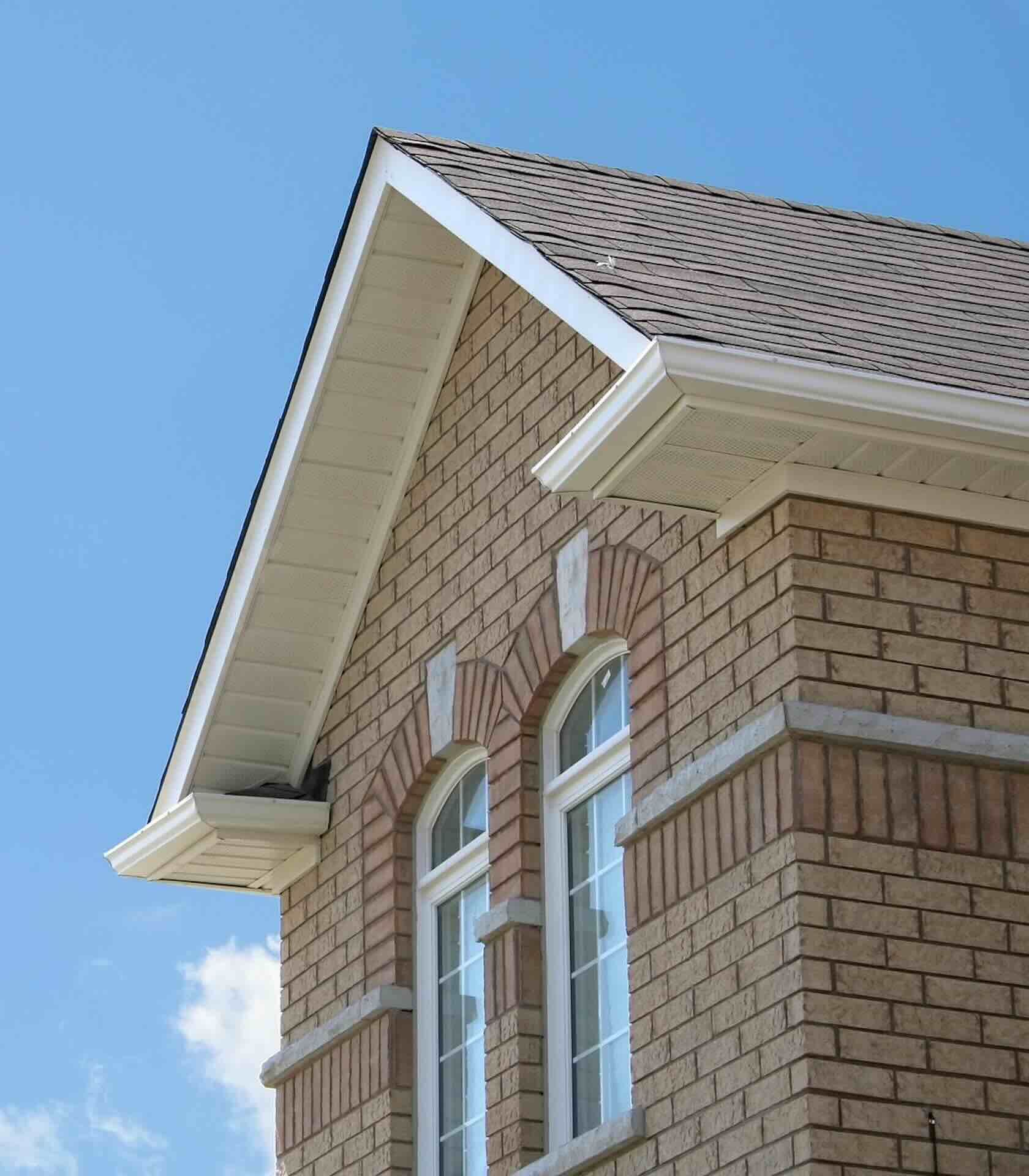 Brick-like siding on house with 2 arched windows under gabled roof, white soffit, and brown shingles against blue sky - Pinnacle Home Improvements