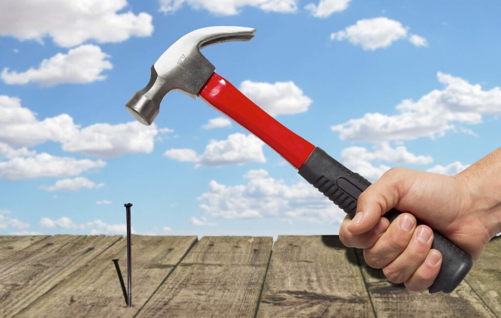 Hand holds red-handled hammer, poised above 2 nails in wooden surface against blue sky with scattered clouds - Pinnacle Home Improvements